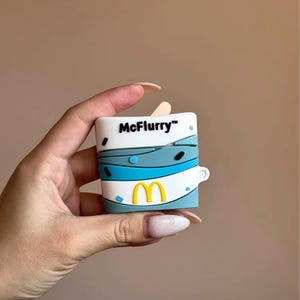 May include: A white and blue McFlurry-themed case for wireless earbuds. The case features the McDonald's logo in yellow and the McFlurry logo in black. The case is held in a hand.