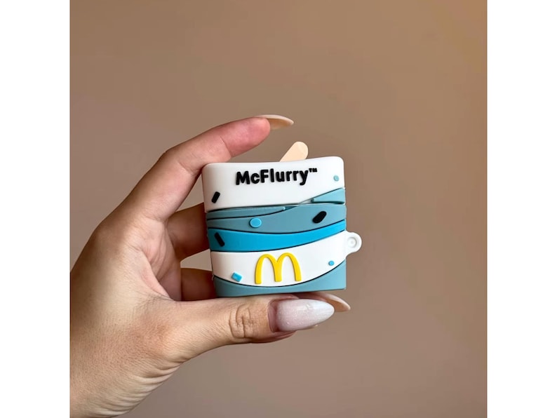 May include: A white and blue McFlurry-themed case for wireless earbuds. The case features the McDonald's logo in yellow and the McFlurry logo in black. The case is held in a hand.