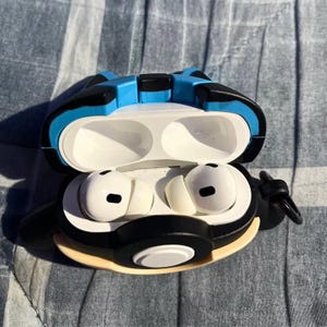 May include: White wireless earbuds inside a black, blue, and tan protective case. The case is open, revealing the earbuds. The case has a cartoon-like design. The background is a blue and white patterned fabric.