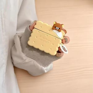 May include: A yellow biscuit-shaped case for wireless earbuds, featuring a brown bear character on top. The case has the words "Rilakkuma BISCUIT" printed on it. A small silver charm hangs from the case.