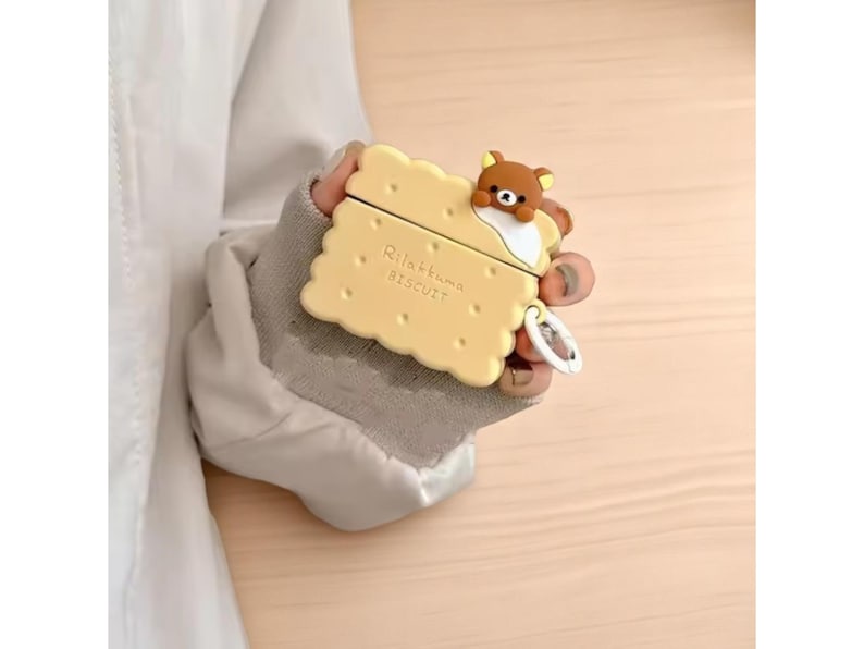 May include: A yellow biscuit-shaped case for wireless earbuds, featuring a brown bear character on top. The case has the words "Rilakkuma BISCUIT" printed on it. A small silver charm hangs from the case.
