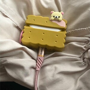 May include: A yellow, biscuit-shaped case with a cartoon bear on top, holding a pink accent. A pink and white Otterbox charging cable is plugged into the case. The case has a playful, whimsical design.