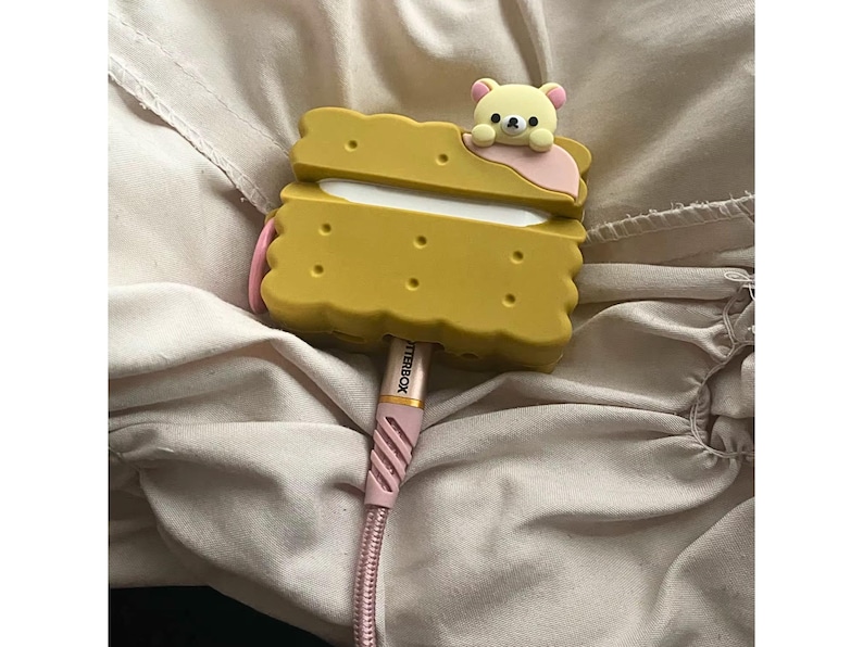 May include: A yellow, biscuit-shaped case with a cartoon bear on top, holding a pink accent. A pink and white Otterbox charging cable is plugged into the case. The case has a playful, whimsical design.
