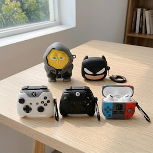 May include: A collection of colorful AirPod cases in various designs. Includes a SpongeBob SquarePants case, a Batman case, two cases shaped like game controllers, and a Nintendo Switch-themed case. The cases are on a light-colored wooden surface.