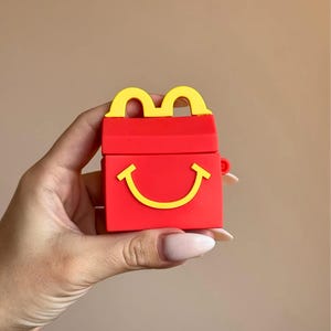 May include: Red and yellow Happy Meal-themed AirPod case. The case is red with a yellow handle and a yellow smiling face. The case is held in a hand against a neutral background.