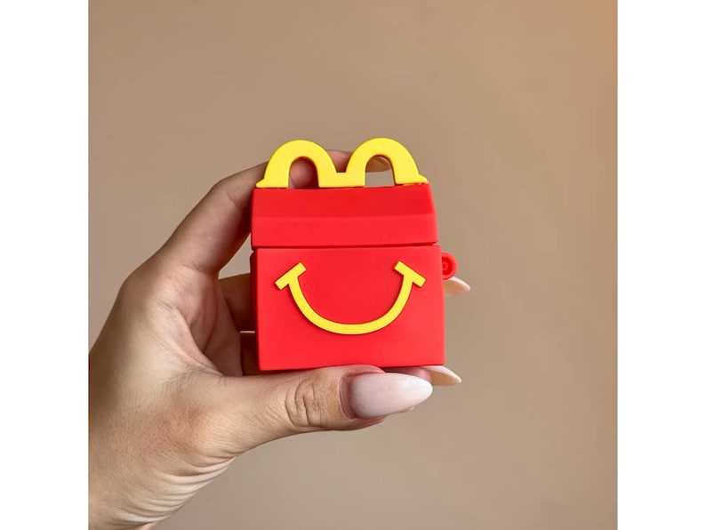 May include: Red and yellow Happy Meal-themed AirPod case. The case is red with a yellow handle and a yellow smiling face. The case is held in a hand against a neutral background.