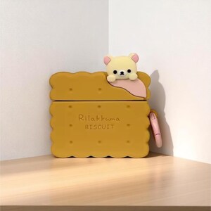 May include: A yellow AirPod case shaped like a biscuit, with the text "Rilakkuma BISCUIT" printed on the front. A small, cream-colored bear figure with pink accents sits on top. A pink strap is attached to the side.