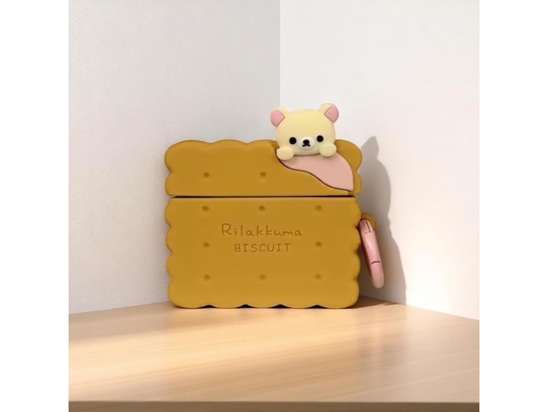 May include: A yellow AirPod case shaped like a biscuit, with the text "Rilakkuma BISCUIT" printed on the front. A small, cream-colored bear figure with pink accents sits on top. A pink strap is attached to the side.