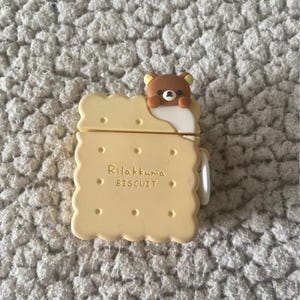 May include: A light yellow AirPod case shaped like a biscuit, with the text "Rilakkuma BISCUIT" printed on the front. A brown bear character peeks over the top edge. The case has a white hinge and is set against a textured, light gray surface.