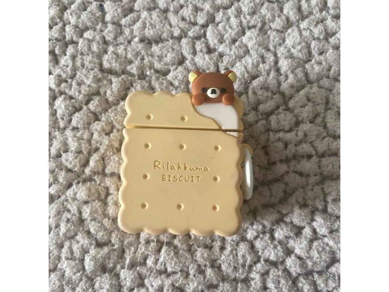 May include: A light yellow AirPod case shaped like a biscuit, with the text "Rilakkuma BISCUIT" printed on the front. A brown bear character peeks over the top edge. The case has a white hinge and is set against a textured, light gray surface.