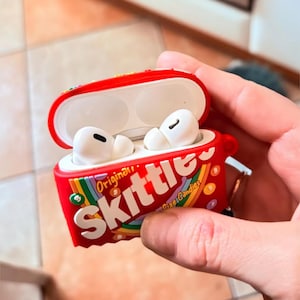 May include: Red and white Skittles-themed AirPod case with white AirPods inside. The case has the Skittles logo and rainbow accents. The case is open, revealing the white earbuds. A keychain is attached to the side.