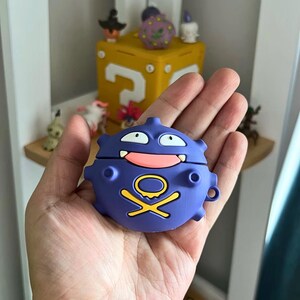 May include: A purple, cartoon-style AirPods case shaped like a Koffing character. The case features white eyes, a pink mouth, and a yellow and black skull and crossbones design. The case is held in a hand.