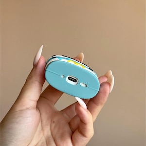 May include: A light blue protective case for wireless earbuds. The case has a colorful design on the top and a charging port on the bottom. The case is held in a hand.