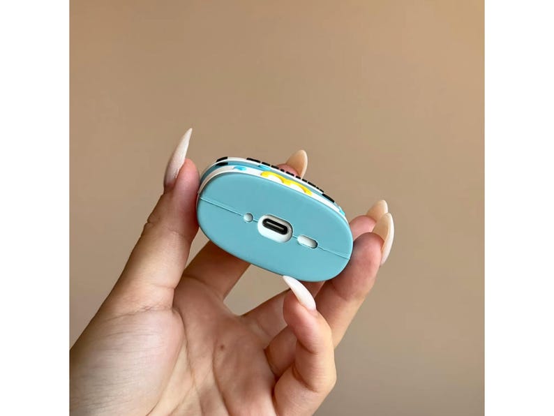May include: A light blue protective case for wireless earbuds. The case has a colorful design on the top and a charging port on the bottom. The case is held in a hand.