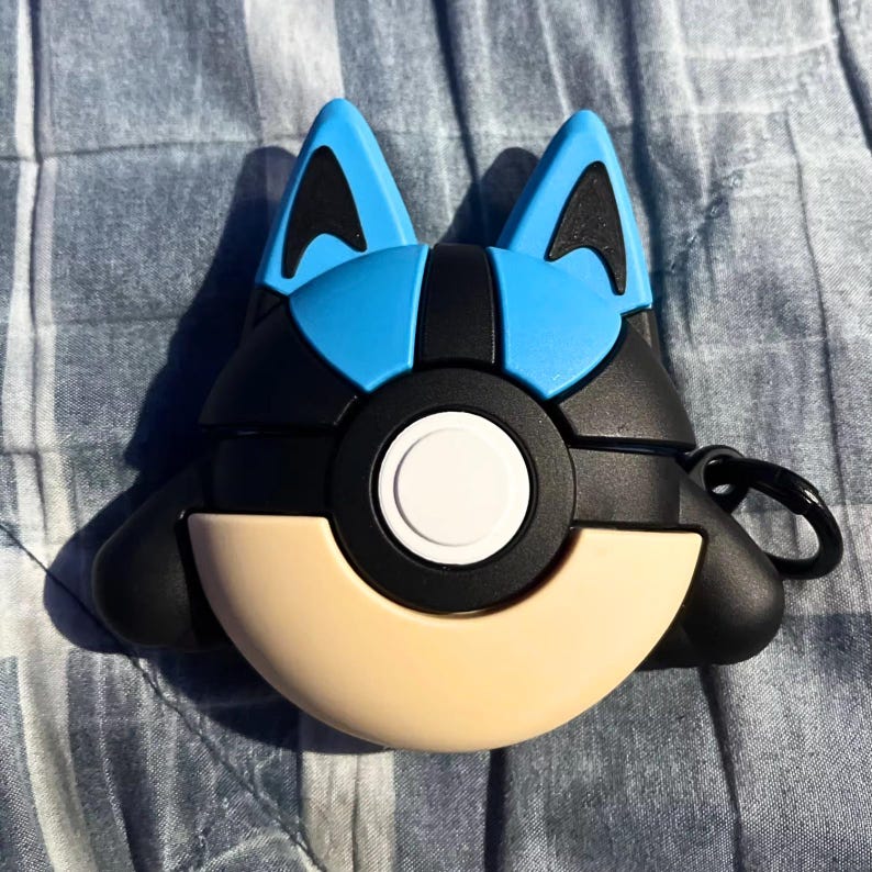 May include: A black, blue, and tan protective case for wireless earbuds. The case is shaped like a cartoon character with blue ears, a black face, and a tan lower section. A black ring is attached for easy carrying.