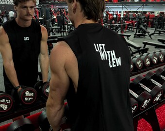 Può includere: Una canotta nera da allenamento con la scritta bianca "LIFT WITH LEW". L'immagine mostra una persona in palestra, con manubri e attrezzature per l'allenamento visibili sullo sfondo. La maglietta ha uno scollo alto.