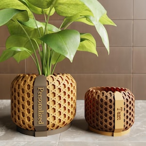May include: Two decorative planters with a woven pattern. The larger planter holds a green plant and has a brown tag that says "Personalized". The smaller planter has a brown tag that says "Text". Both planters are shades of brown.