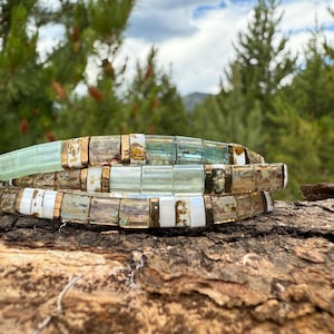 May include: Three stacked bracelets with rectangular beads in shades of white, clear, and pale green, accented with gold-colored metal. The bracelets are displayed on a piece of wood, with a blurred background of green trees and a cloudy sky.