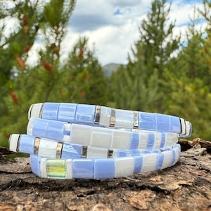 May include: Four stacked bracelets with a mosaic tile design. The bracelets feature alternating light blue and white square tiles, separated by silver-colored metal accents. The bracelets are displayed on a piece of wood, with a blurred green background.