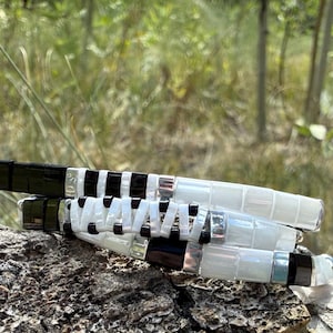 May include: Three stacked bracelets with a mix of black, white, and silver beads. The bracelets are resting on a textured, brown surface, with a blurred green background of foliage and trees.