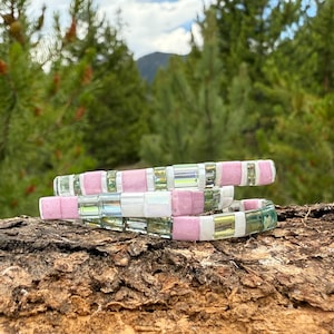May include: Three stacked bracelets with a mix of pink, iridescent, and clear rectangular beads. The bracelets are displayed on a piece of wood, with a blurred background of green trees and a mountain.