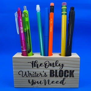 May include: Wooden pencil holder with the text "The Only Writer's Block You Need" engraved on the front. The holder has six slots and is filled with various writing utensils, including a pink pen, a green pen, a blue pen, an orange pencil, a yellow pencil, and a black pen.