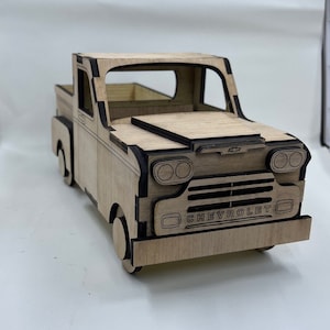 Peut inclure: Maquette en bois d'un pick-up Chevrolet vintage. Le camion est en bois clair avec des contours noirs et le mot "CHEVROLET" à l'avant. Le modèle présente des découpes détaillées pour les fenêtres, les phares et d'autres éléments.