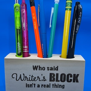 May include: Wooden pencil holder with the text "Who said Writer's BLOCK isn't a real thing" engraved on the front. The holder has six slots and is filled with various writing utensils, including a green pen, a pink pen, an orange pencil, a blue pen, a green pen, a yellow pencil, and a black pen.
