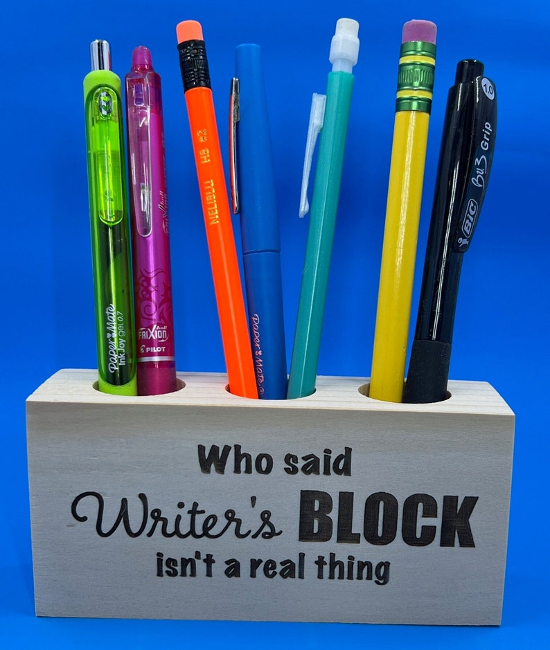 May include: Wooden pencil holder with the text "Who said Writer's BLOCK isn't a real thing" engraved on the front. The holder has six slots and is filled with various writing utensils, including a green pen, a pink pen, an orange pencil, a blue pen, a green pen, a yellow pencil, and a black pen.