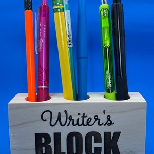 May include: Wooden pencil holder with the words "Writer's Block" engraved on the front. The holder has five slots and is filled with various writing utensils, including pencils, pens, and highlighters.