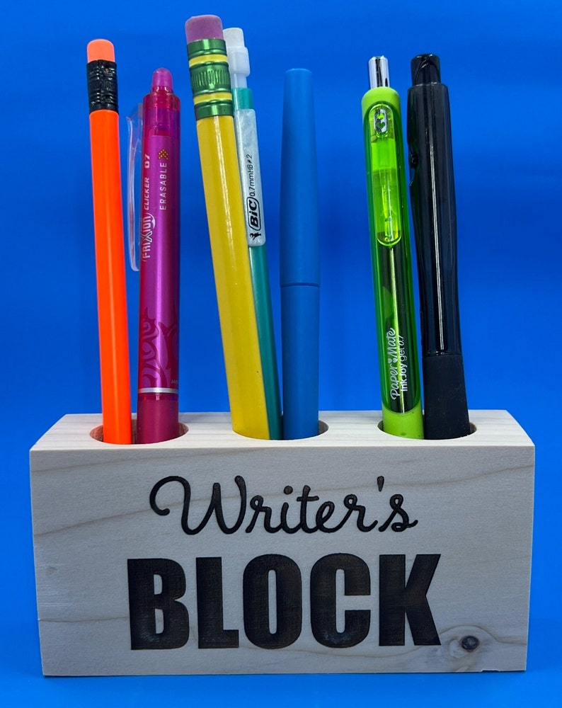 May include: Wooden pencil holder with the words "Writer's Block" engraved on the front. The holder has five slots and is filled with various writing utensils, including pencils, pens, and highlighters.