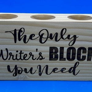 May include: A wooden pencil holder with three holes. The holder is light brown and has the text "The Only Writer's Block You Need" engraved on the front.