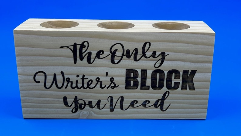 May include: A wooden pencil holder with three holes. The holder is light brown and has the text "The Only Writer's Block You Need" engraved on the front.