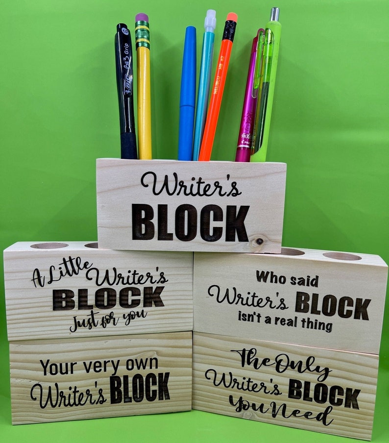 May include: Four wooden pencil holders with different sayings about writer's block. The top holder says "Writer's Block". The second holder says "A Little Writer's Block Just for You". The third holder says "Who said Writer's Block isn't a real thing". The bottom holder says "The Only Writer's Block You Need".