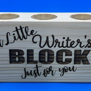 May include: A wooden pencil holder with three slots. It is engraved with the text "A little Writer's BLOCK Just for you".