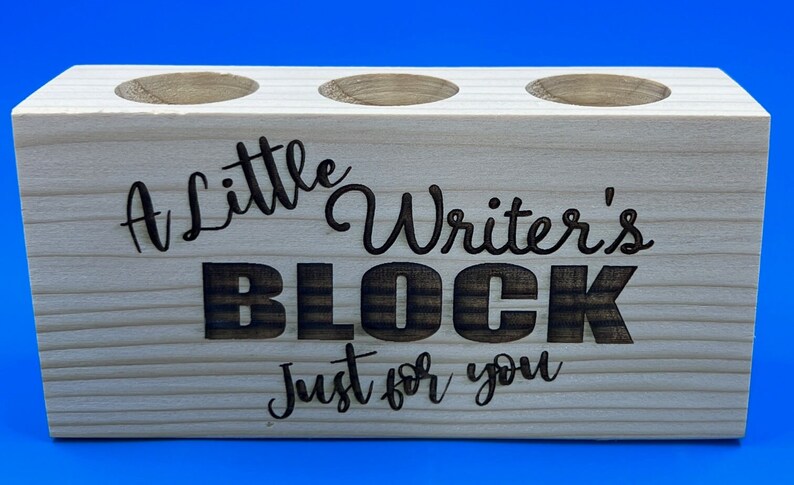May include: A wooden pencil holder with three slots. It is engraved with the text "A little Writer's BLOCK Just for you".