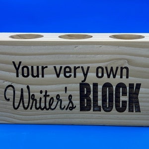 May include: A wooden pencil holder with three slots. The holder is engraved with the text "Your very own Writer's BLOCK".