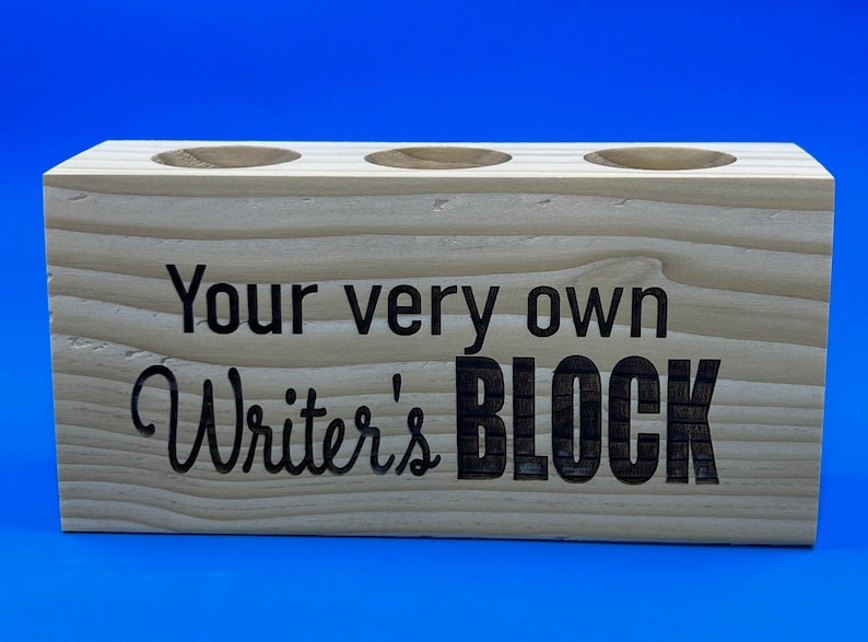 May include: A wooden pencil holder with three slots. The holder is engraved with the text "Your very own Writer's BLOCK".