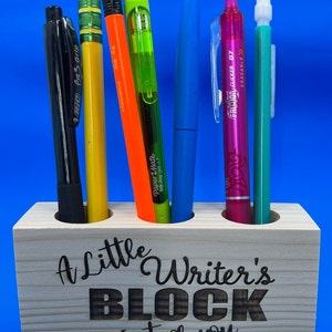 May include: A wooden pencil holder with the text "A Little Writer's Block Just for You" engraved on the front. The holder has three slots and is filled with various writing utensils, including a black pen, a yellow pencil, an orange pen, a green pen, a blue pen, a pink pen, and a turquoise pen.