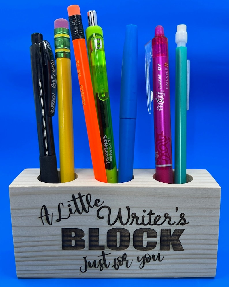 May include: A wooden pencil holder with the text "A Little Writer's Block Just for You" engraved on the front. The holder has three slots and is filled with various writing utensils, including a black pen, a yellow pencil, an orange pen, a green pen, a blue pen, a pink pen, and a turquoise pen.