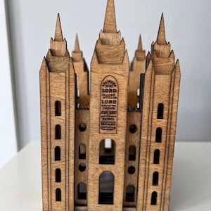 Op de afbeelding: Een houten model van een kerk met een toren en meerdere torens. De kerk is gemaakt van lichtbruin hout en heeft een uitgesneden ontwerp. De tekst "HOLINESS TO THE LORD. HOUSE OF THE LORD" is gegraveerd op de voorkant van de kerk.