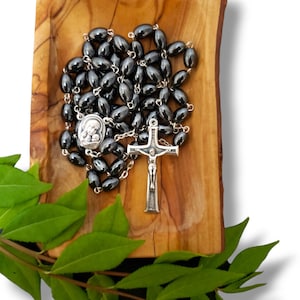 May include: A black rosary with oval beads and a silver crucifix. The rosary is displayed on a wooden surface with green leaves in the foreground. The crucifix features a detailed depiction of Jesus Christ.