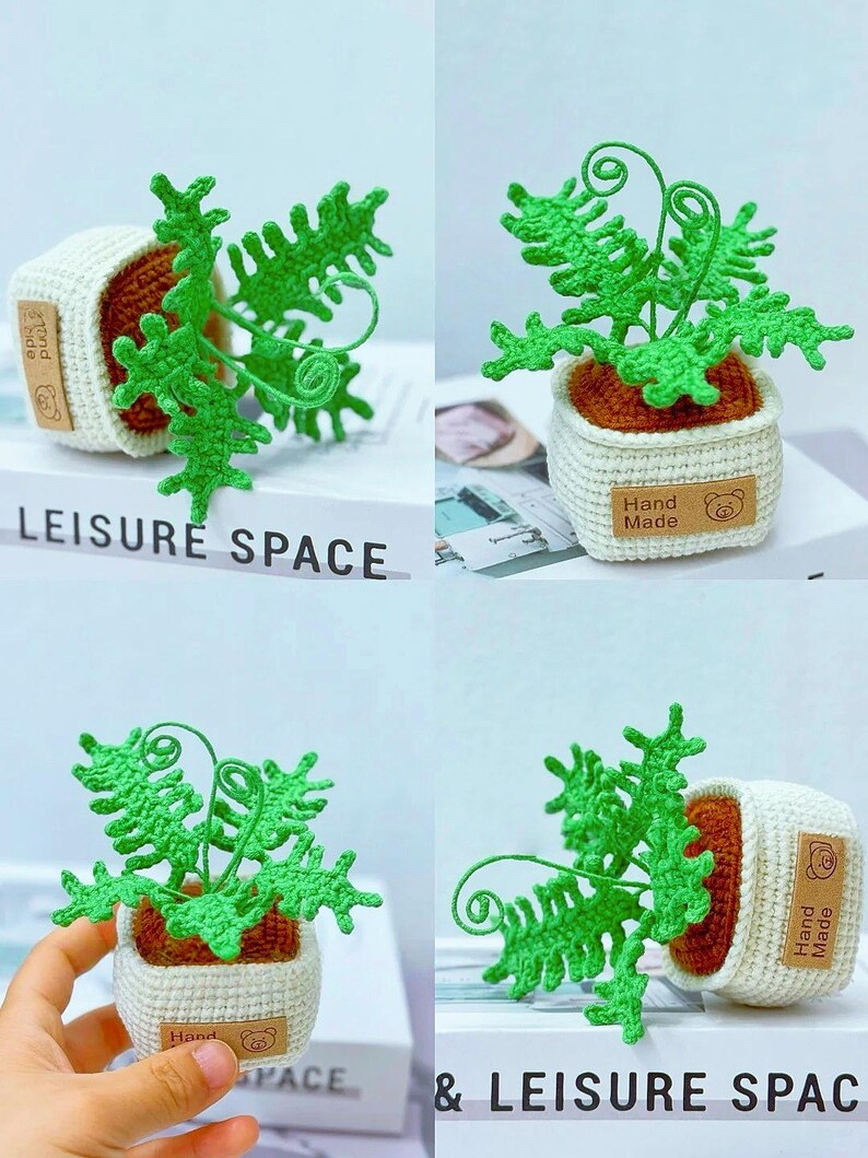 K&ouml;nnte beinhalten: Geh&auml;kelte Miniatur-Farnpflanze in einem wei&szlig;en Topf mit braunem Innenraum. Der Farn ist hellgr&uuml;n, und der Topf hat ein "Hand Made"-Etikett mit einem B&auml;ren. Der Text "LEISURE SPACE" ist sichtbar. Ein charmantes handgefertigtes Dekorationsst&uuml;ck.