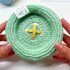May include: A round, light green crocheted bowl with a thick, raised rim. The center features a yellow crocheted cross design resembling a button. A white fabric tag with black text and a small line drawing is attached to the inner rim.