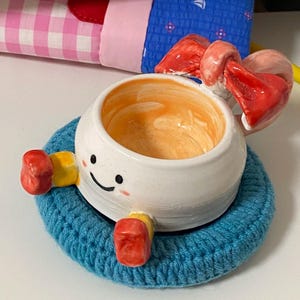May include: A white ceramic bowl with a hand-painted smiling face, orange interior, and red bow detail. The bowl features small yellow and red limbs and rests on a circular blue crocheted coaster. The design includes a glossy finish.