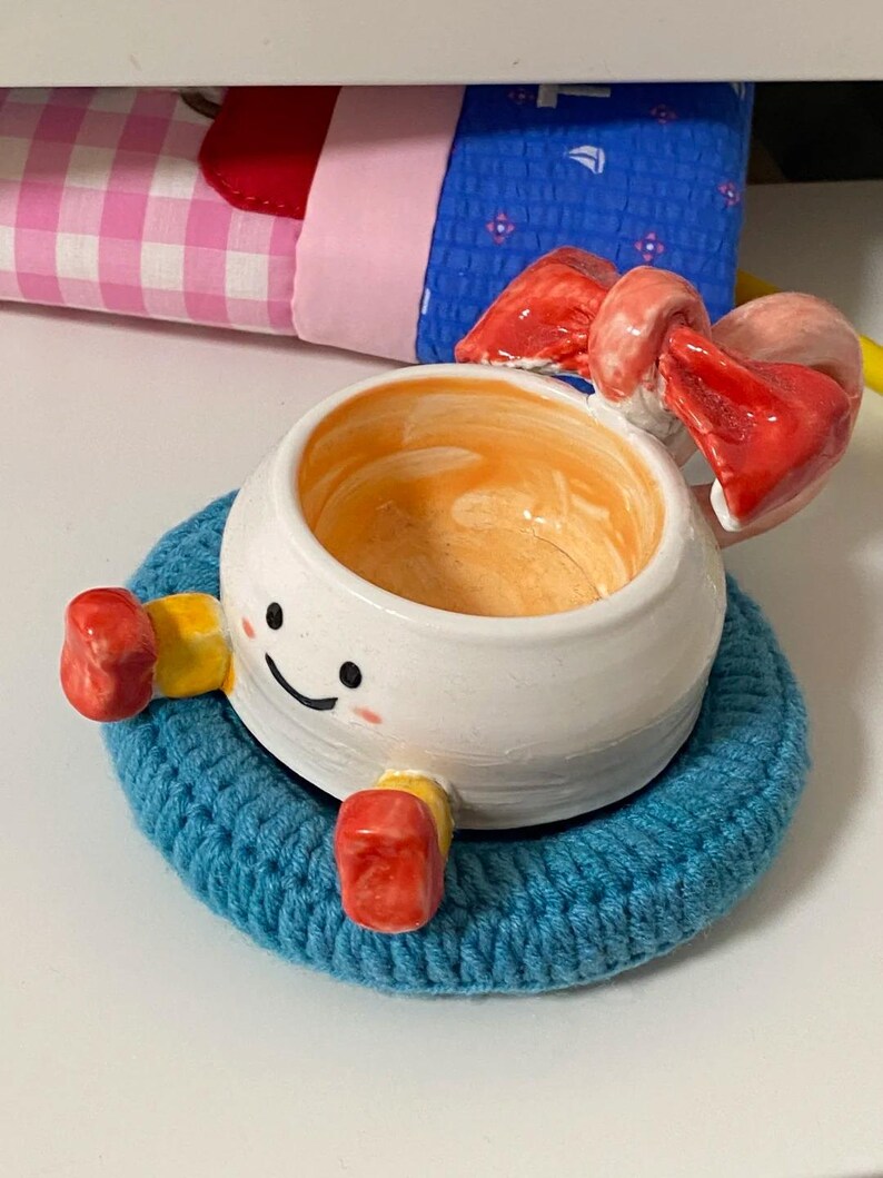 May include: A white ceramic bowl with a hand-painted smiling face, orange interior, and red bow detail. The bowl features small yellow and red limbs and rests on a circular blue crocheted coaster. The design includes a glossy finish.
