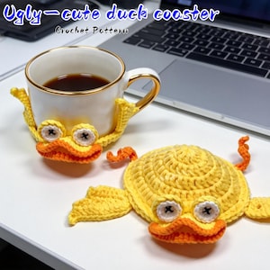 Könnte beinhalten: Eine weiße Keramiktasse mit goldenem Rand, gefüllt mit Kaffee, steht auf einem gelb-orangen gehäkelten Entenuntersetzer. Ein zweiter Entenuntersetzer ist ebenfalls sichtbar. Der Text "Ugly-cute duck coaster" und "Crochet Pattern" stehen oben.
