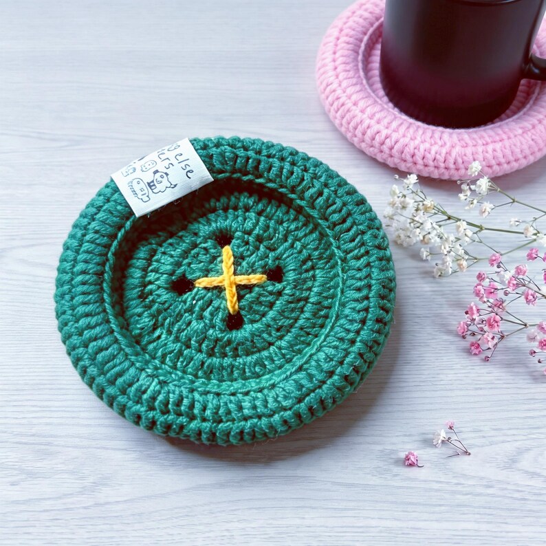 May include: A round, dark green crocheted coaster shaped like a large button with a yellow cross-stitched center. A small white fabric tag with black line art and text is attached to the edge. The coaster features a thick, textured yarn border.