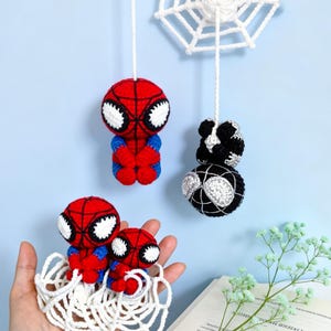 Puede incluir: Una colección de figuras de ganchillo con personajes rojos y azules con grandes ojos blancos y patrones de telaraña negros, junto a un personaje en blanco y negro, todos suspendidos por hilos de lana blancos a una telaraña de ganchillo.