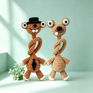 May include: Two handcrafted crochet dolls with quirky features. One doll is adorned with a black top hat and a black tie, the other is plain. Both dolls have large, round eyes and twisted necks. The dolls are brown and tan with white detailing.